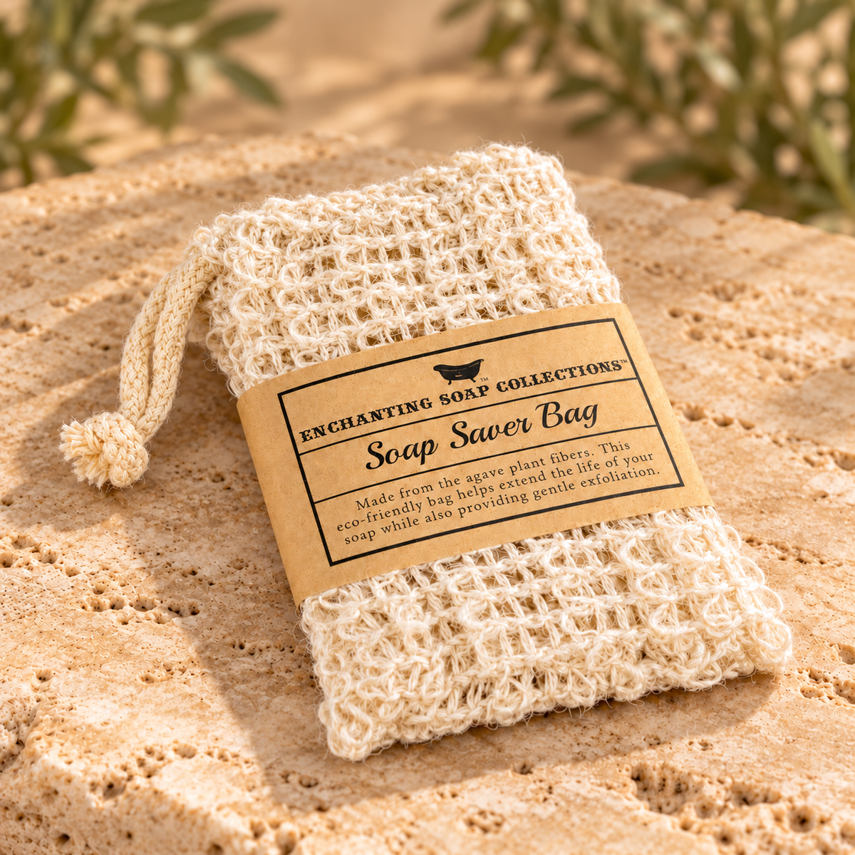 A natural beige soap saver bag made from agate plant fibers, wrapped in a kraft paper label that reads 'Enchanting Soap Collections Soap Saver Bag,' resting on a sandy surface with blurred greenery in the background.